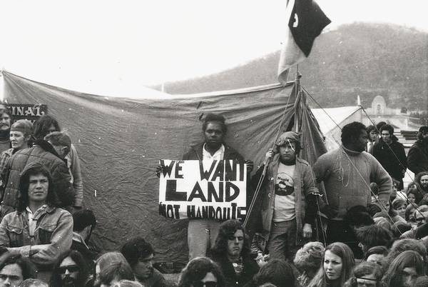 A large group of people, mostly sitting, in front of a tent. In the middle of the image Alan Sharpley stands holding a sign with black lettering on a white background: We want land rights not handouts. A flag flies from the front of the tent and the Australian War Memorial can be seen far in the background.
